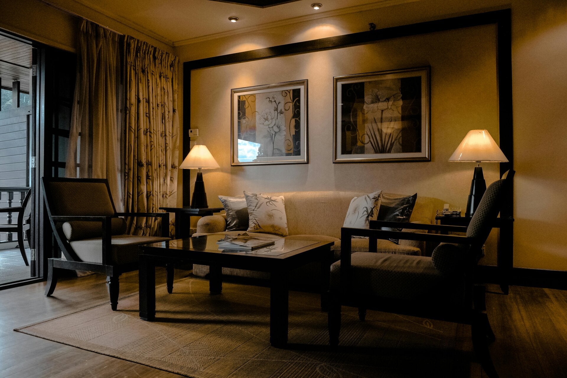 Warmly lit traditional living room in the evening with a beige sofa, dark wood armchairs, a black lacquer coffee table, matching table lamps and framed floral prints